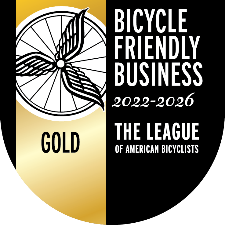 Badge stating Gold Bicycle Friendly Business 2022-2026 from The League of American Bicyclists. It has a gold stripe plus an image of a bicycle wheel that has three pretend "wings" on it.