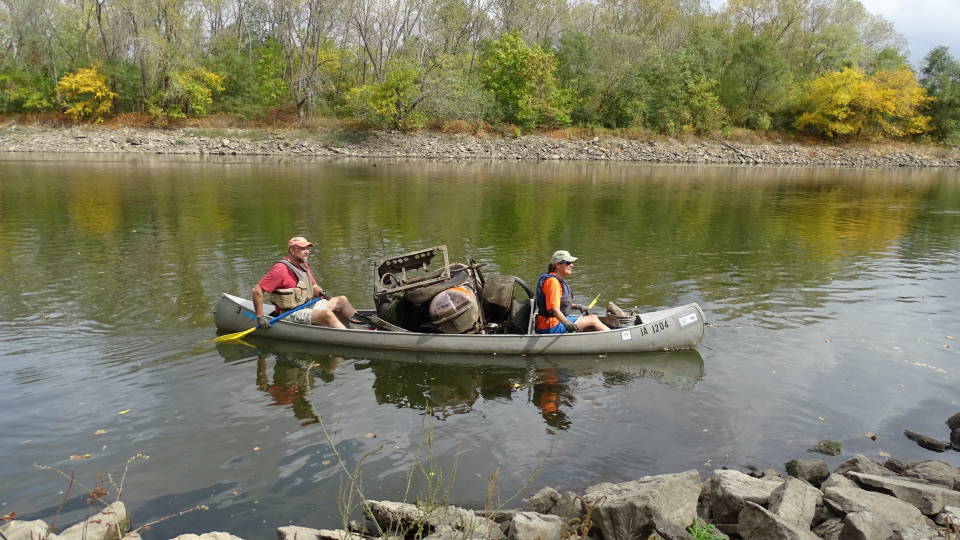 A canoe loaded with large pieces of trash, including barrels, tires, and miscellaneous junk metal, is paddled down the river by two adults.