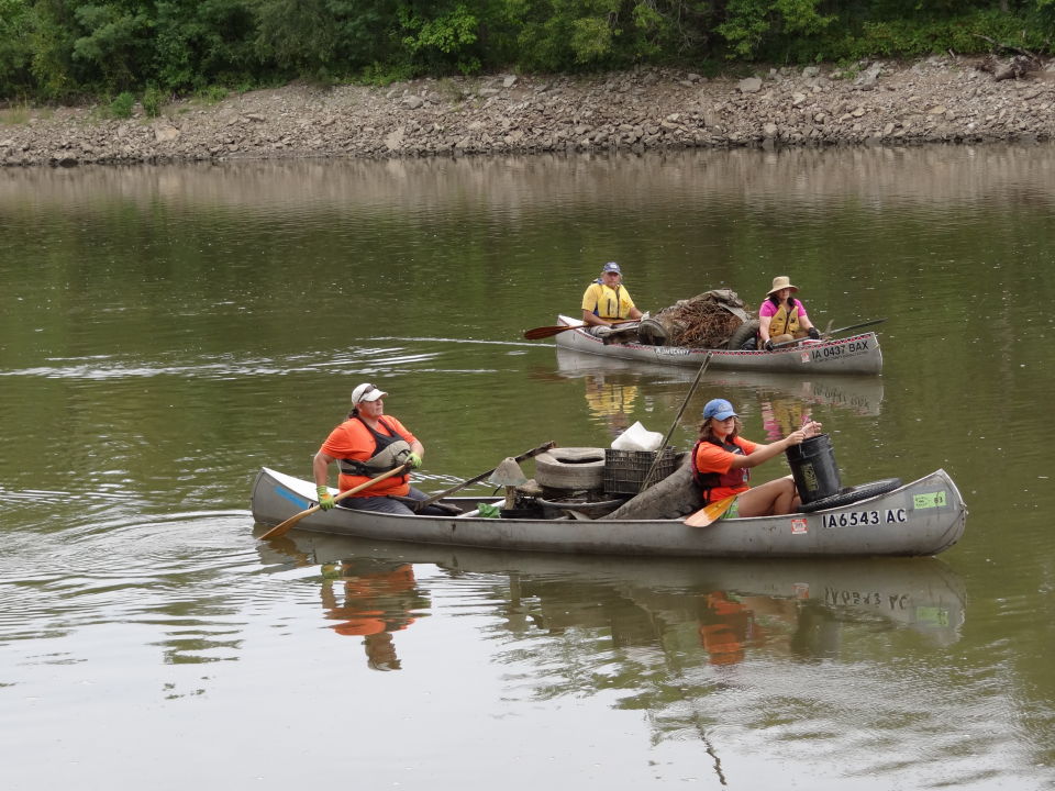 Two canoes loaded with trash such as tires, barbed wire, and crates are paddled by two adults and two kids.