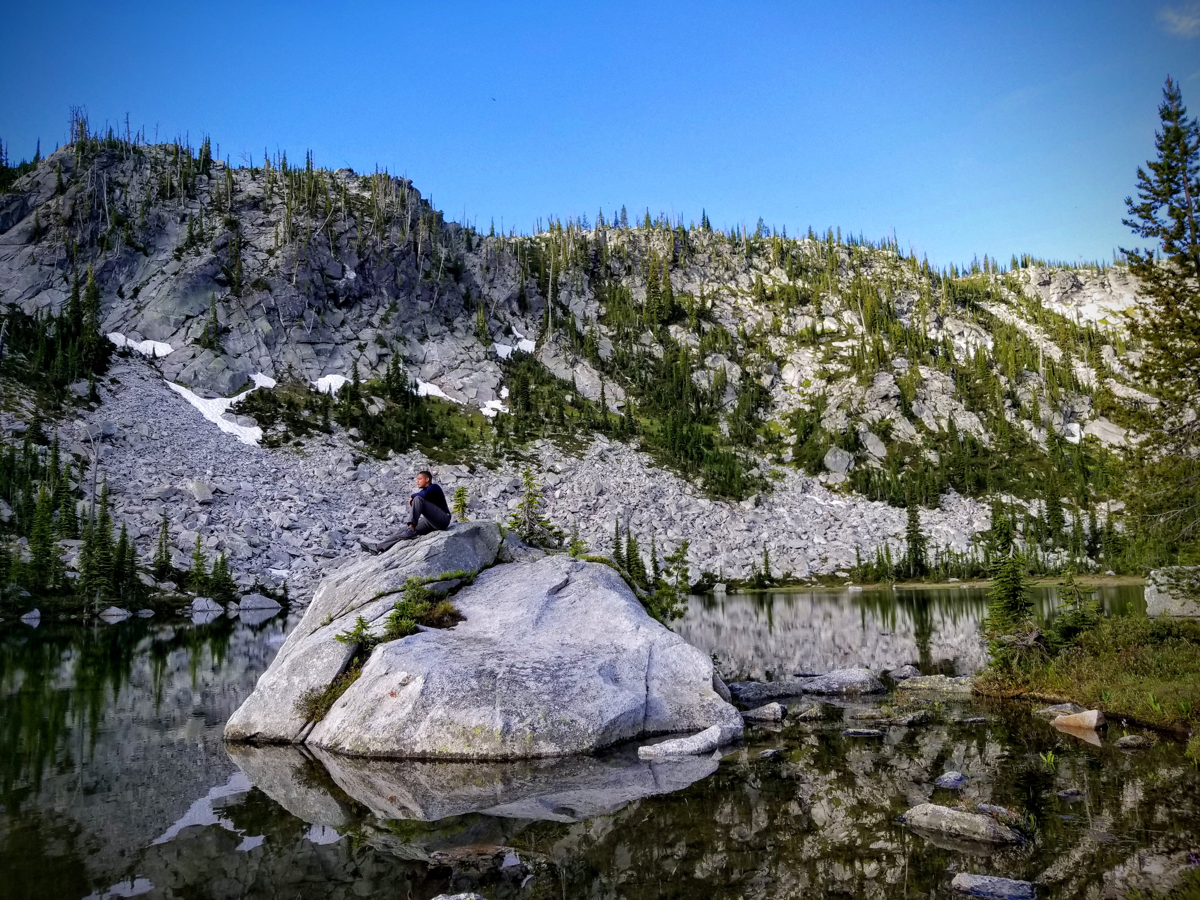 A high school boy sits on a large boulder surrounded by a lake and mountains.  