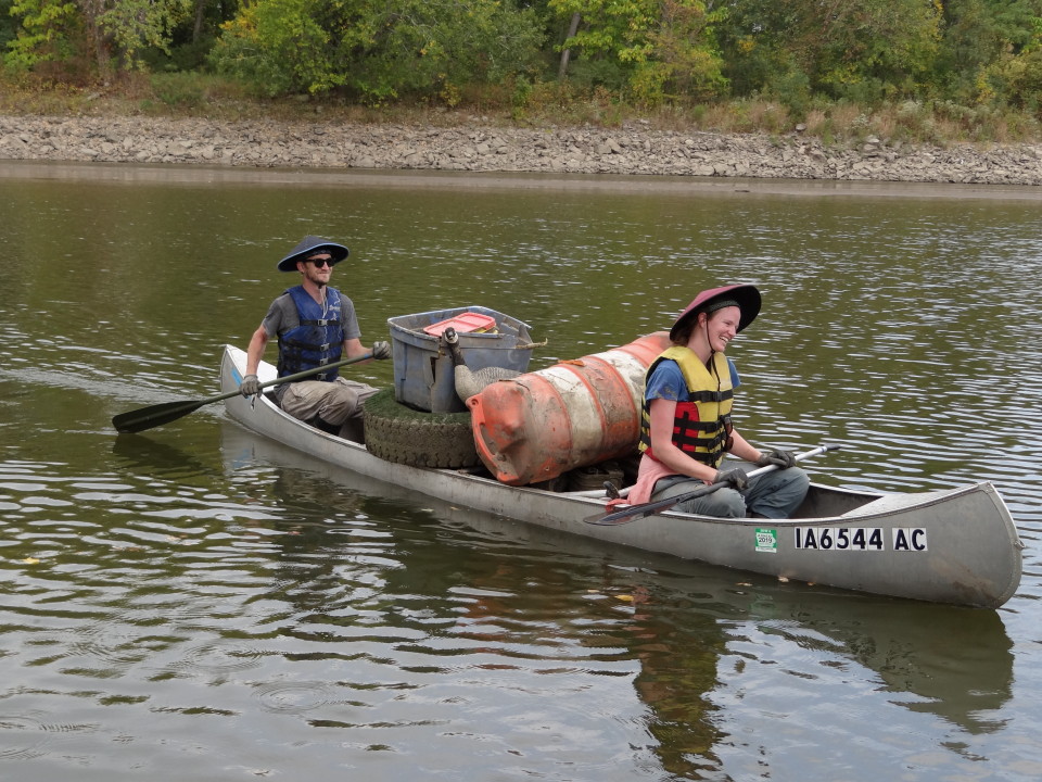 A man and woman paddle a canoe that is loaded with trash such as a tire, large traffic cone, goose decoy, and a large plastic tub. 