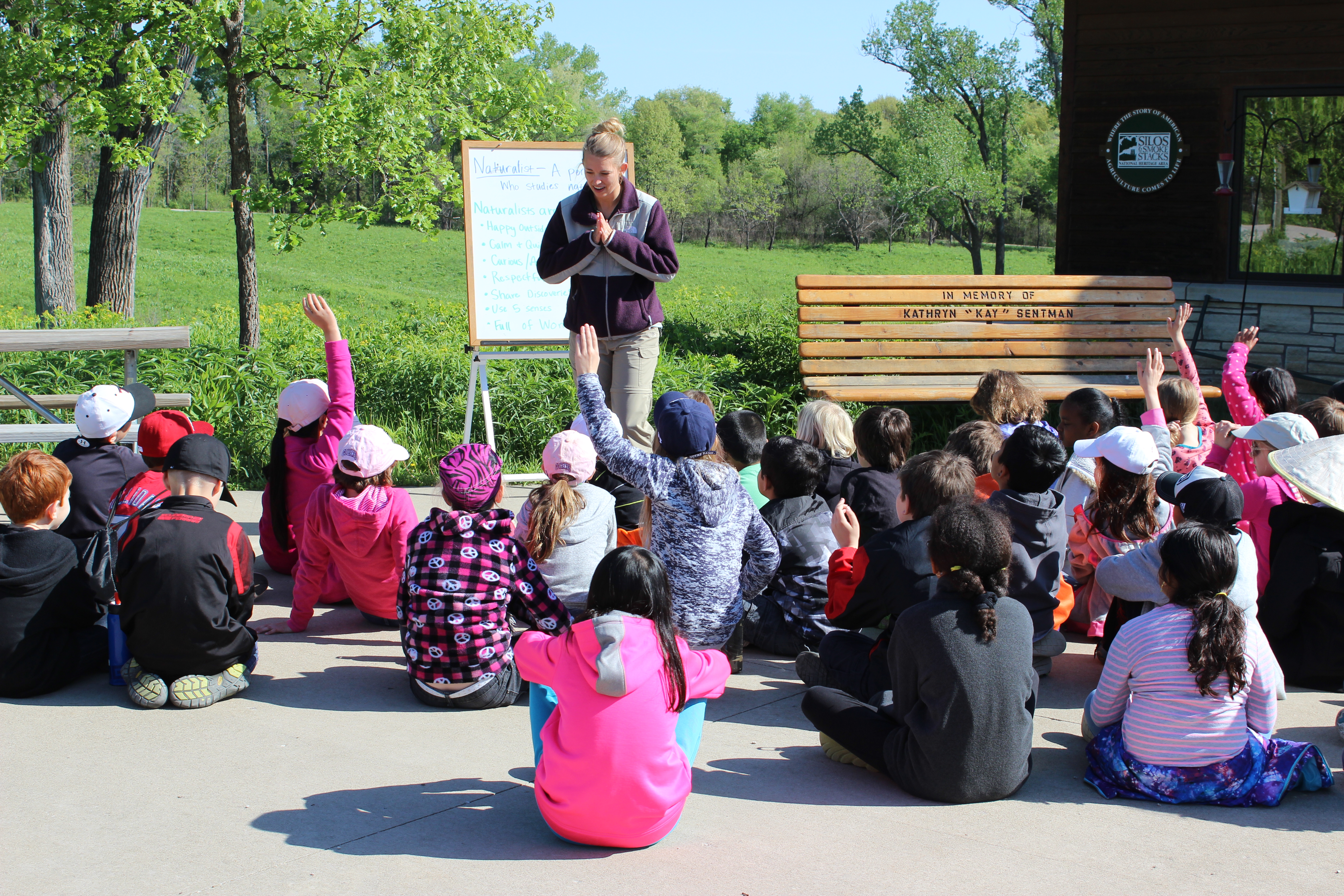 A naturalist talks to a large group of students while standing in front of a white board outdoors.